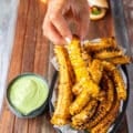 A hand holding a corn rib cooked and ready, with a side of avocado crema