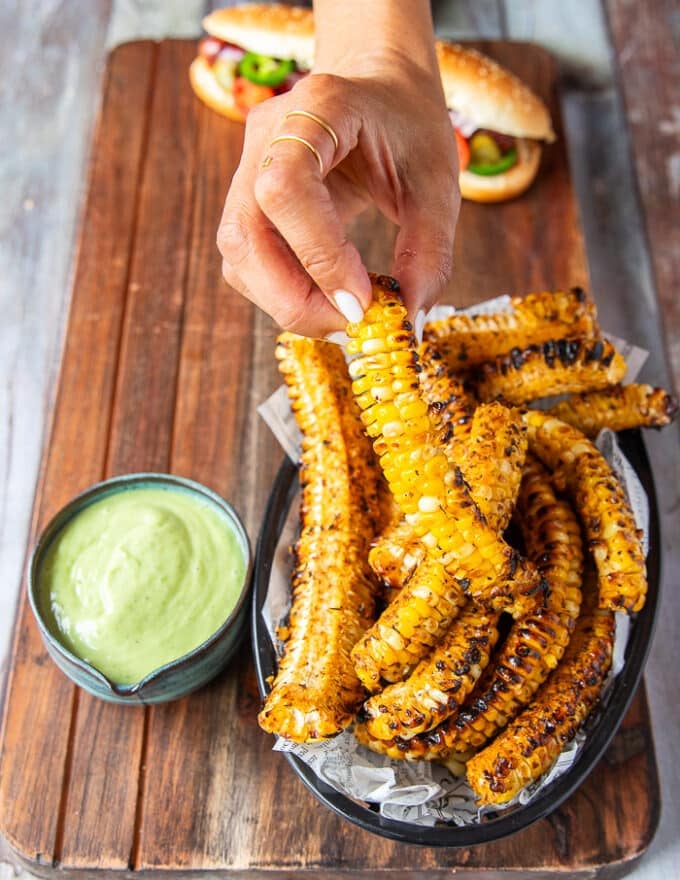 A hand holding a corn rib cooked and ready, with a side of avocado crema 