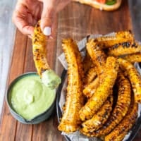 A hand holding a piece of seasoned corn ribs dipped into the avocado jalapeno sauce and showing close up of the dip