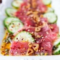close up shot showing the tuna thinly sliced and sititng in the vinaigrette with a slice of cucumber pn the side, some garlic chips and capers around it. The texture of the tuna crudo is showing so clealry