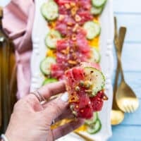 A hand holding a piece of tuna, cucumber over a crunchy Italian ciabbata bread showing how to serve tuna crudo with bread