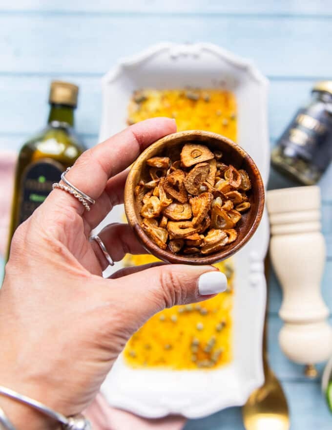A hand holding a small bowl of golden toasted garlic chips ready to garnish the tuna crudo 