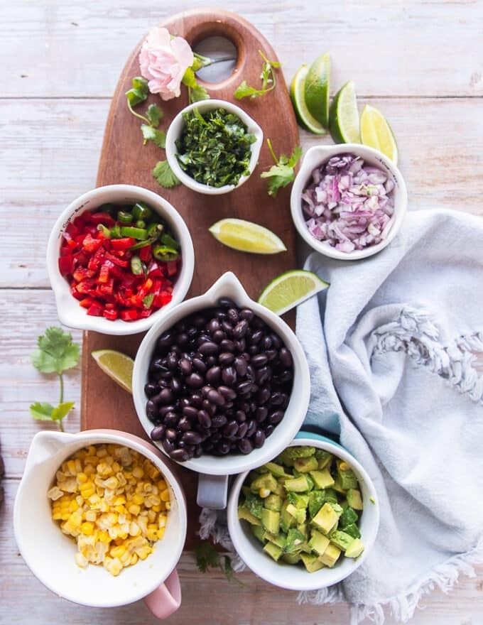 Ingredients for the black bean salad including a bowl of black bean, a bowl of fresh corn, some diced avocado, some diced onions in a bowl, some red peppers, a bowl of fresh minced cilantro, some lime on a board