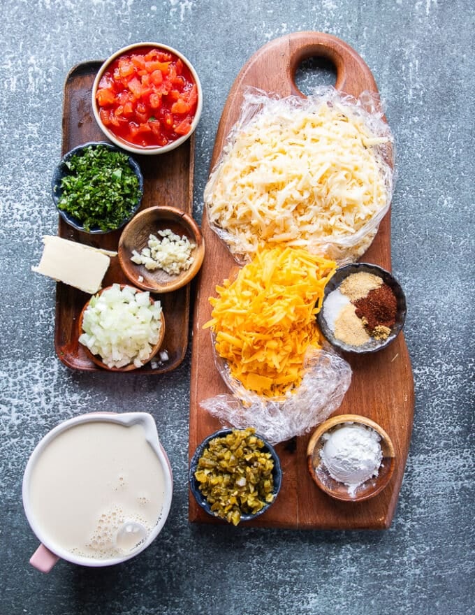 Ingredients in small bowls on a wooden board including shredded cheeses (cheddar and Monterey jack) some cornstarch, some diced onions, minced garlic, minced jalapeño, diced tomatoes, a bowl of spices, butter, pickled jalapeños, cilantro and a glass of milk.