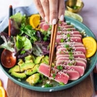 A hand holding one piece of tuna tataki with chopsticks up close showing the texture and ponzu dressing on top