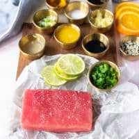 tuna tataki ingredients including the ahi tuna on a board surrounded by lime juice in a cup, some orange juice in another cup, ginger grated in a cup, garlic, scallion minced, sesame seeds in a bowl, sugar in a bowl, soy sauce in a bowl, cilantro in a bowl