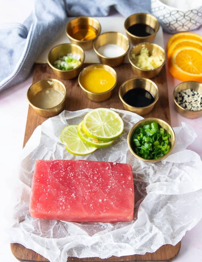 tuna tataki ingredients including the ahi tuna on a board surrounded by lime juice in a cup, some orange juice in another cup, ginger grated in a cup, garlic, scallion minced, sesame seeds in a bowl, sugar in a bowl, soy sauce in a bowl, cilantro in a bowl