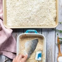 A hand dipping a piece of perch fish into the wet ingredients plate