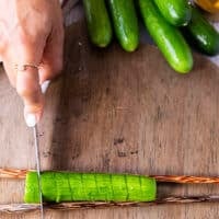 The cucumber is flipped and still over the chopsticks, a hand making straight cuts with no angle on the cucumbers using the same knife