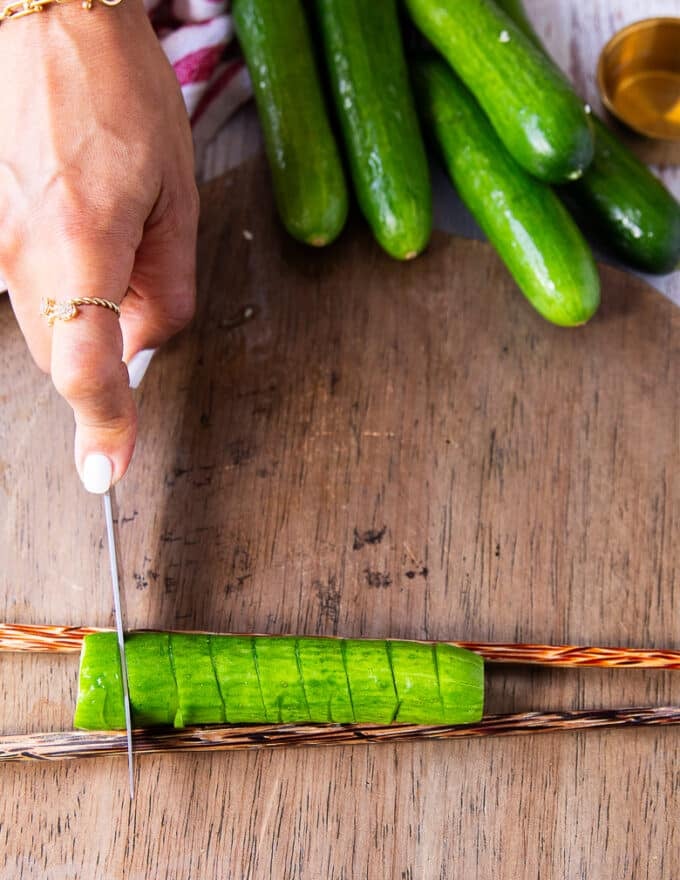 The cucumber is flipped and still over the chopsticks, a hand making straight cuts with no angle on the cucumbers using the same knife