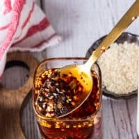 Close up of the chili oil recipe when mixed with a spoon showing the red hot oil infused with the chilli flakes and the bits of chillies, sesame , scallions, ginger, garlic within the oil