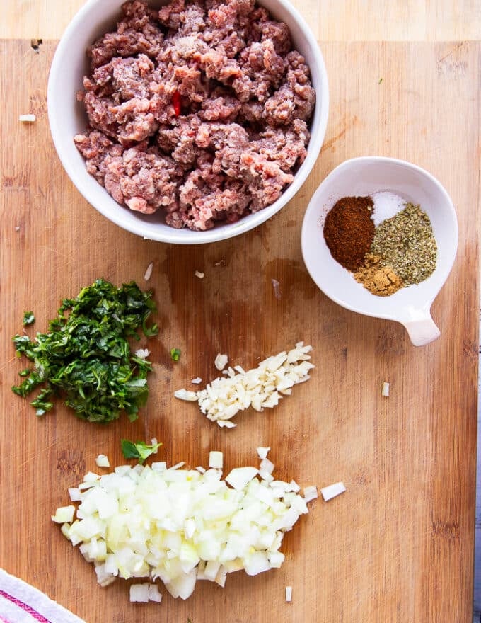 ingredients for ground beef nachos including the beef, diced onions, garlic, cilantro and a homemade taco mix on a cutting board, some chips, nacho cheese sauce, salsa, avocado, guacamole and sour cream