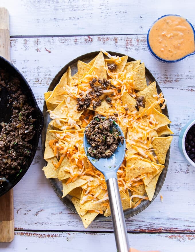 The chips are layered at the bottom of the serving dish, some shredded cheese and a hand adding in the crumbled ground beef on top 