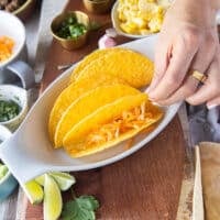 A hand holding a hard taco shell in a baking sheet and adding cheese to the taco shell to start assembling the ground beef tacos