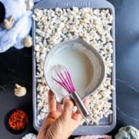A hand holding a bowl of melted white chocolate to pour over the popcorn on a baking sheet