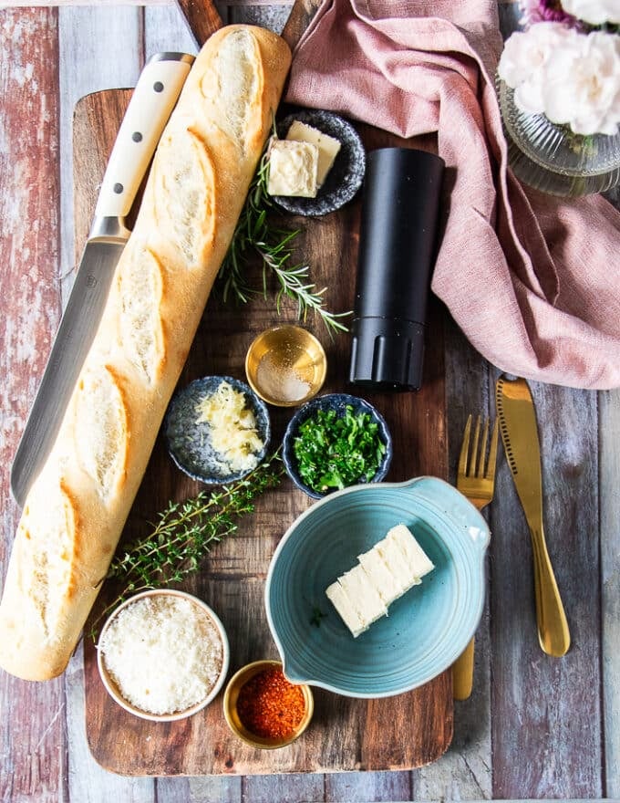 Ingredients for air fryer garlic bread on a borad including a loaf of baguette bread, some butter in a bowl, some fresh herbs, some grated parmesan cheese, some salt, pepper and fresh garlic minced , a bowl of garlic powder and tea towel