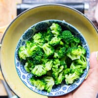 A hand holding a bowl of mini broccoli florets chopped finely to add to the final pot