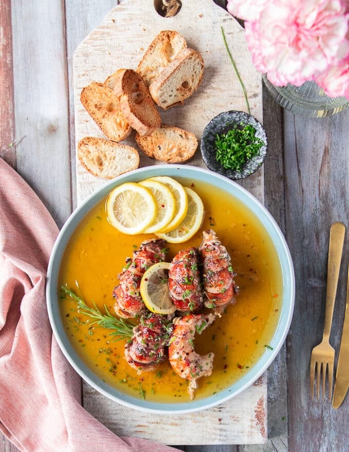 A finished plate of butter poached lobsters drenched in the butter sauce served with bread, some lemon slices, chilli flakes and chopped chives 