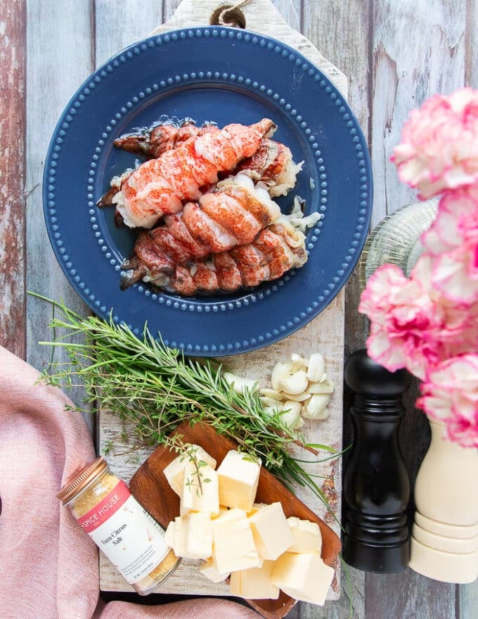 butter poached lobster ingredients on a wooden board including some lobster tails, some diced butter, lots garlic and some rosemary, some seasoning 
