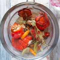 The lobster stock is passed through a strainer showing the lobster shells, all the veggies and herbs in the strainer, and a clear golden liquid left