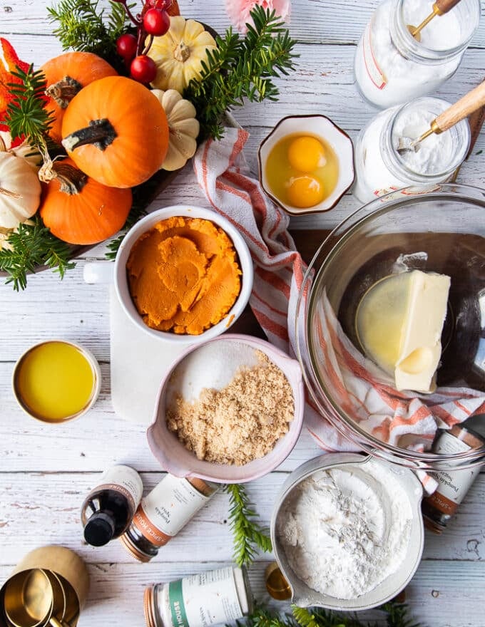 ingredients for pumpkin bread recipe including a bowl of pumpkin pure, butter in a large bowl, some brown sugar in a bowl, white sugar in a bowl, an egg in a bowl, the flour mixture in a separate bowl, vanilla and pumpkin spice 