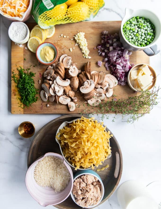 Ingredients on a large marble for tuna casserole. Caned tuna in a bowl, cheese in a bowl, some milk, some butter in separate bowls, the egg noddles uncooked in a bowl, sliced mushrooms on a board with diced onions, some garlic minced, a bowl of dijon mustard, some frozen peas in a bowl,some lemon slices, a bowl of seasoned panko, some half n half in a cup