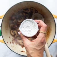 A hand sprinkling flour over the onion and mushrooms