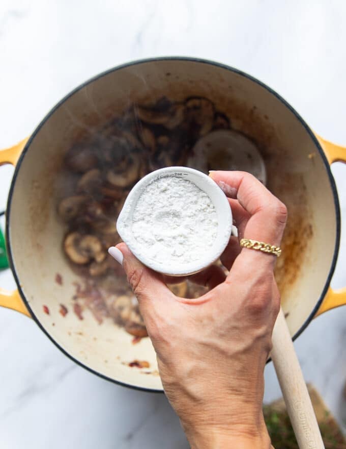 A hand sprinkling flour over the onion and mushrooms 
