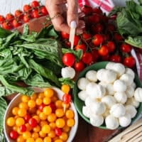 A hand holding a bamboo skewer with a basil leaf skewered followed by cherry tomato, bocconcini , more basil, baby orange and yellow tomatoes and another basil leaf