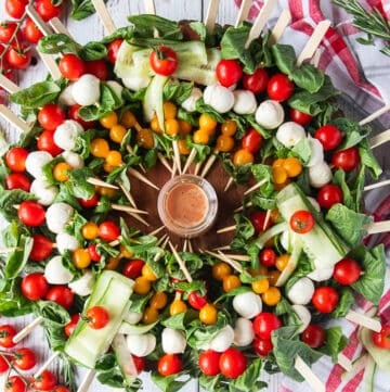 A whole caprese wreath with skewers of tomatoes, basil and mini mozzarella cheese on a bed of basil leaveas and a small bowl of vinaigrette in he middle