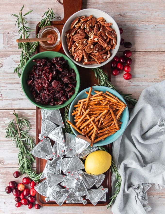 Ingredients to make the cheese Christmas trees on a wooden board including cheese wedges, pretzel sticks, a bowl of cranberries, a bowl of pecans, a lemon and lots of fresh rosemary, and a jar of honey
