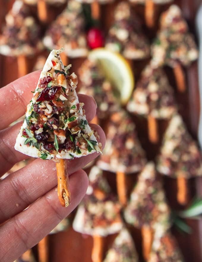 A hand holding one piece of Christmas cheese tree over the board of cheese appetizer and showing close up the seasoning mix, the pretzel stick and the perfect tree shape