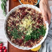 A hand holding aa shallow plate with crushed pecans, cranberries, lemon zest, fresh rosemary, parsley , honey and seasoning.