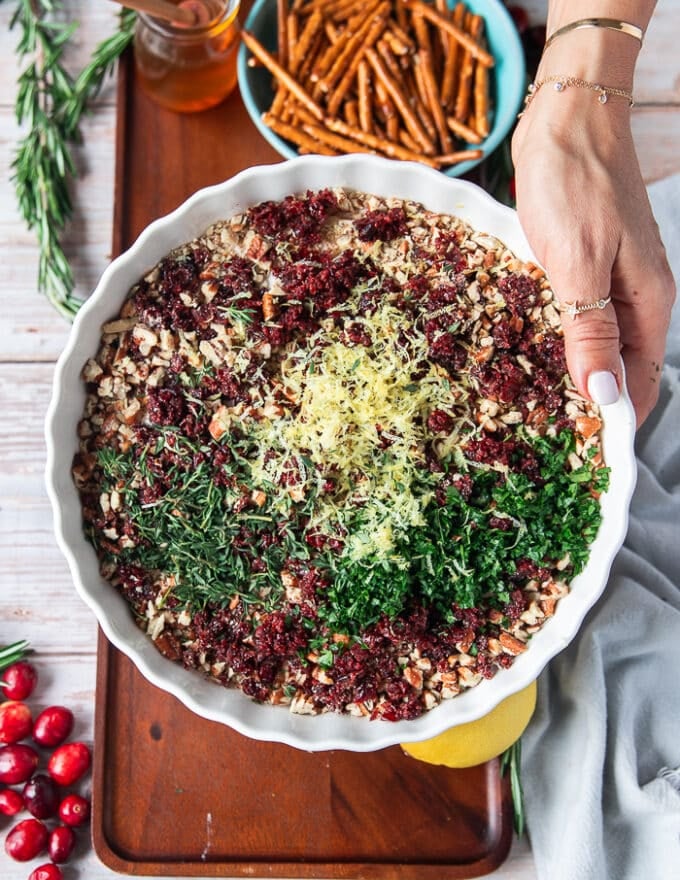 A hand holding aa shallow plate with crushed pecans, cranberries, lemon zest, fresh rosemary, parsley , honey and seasoning.