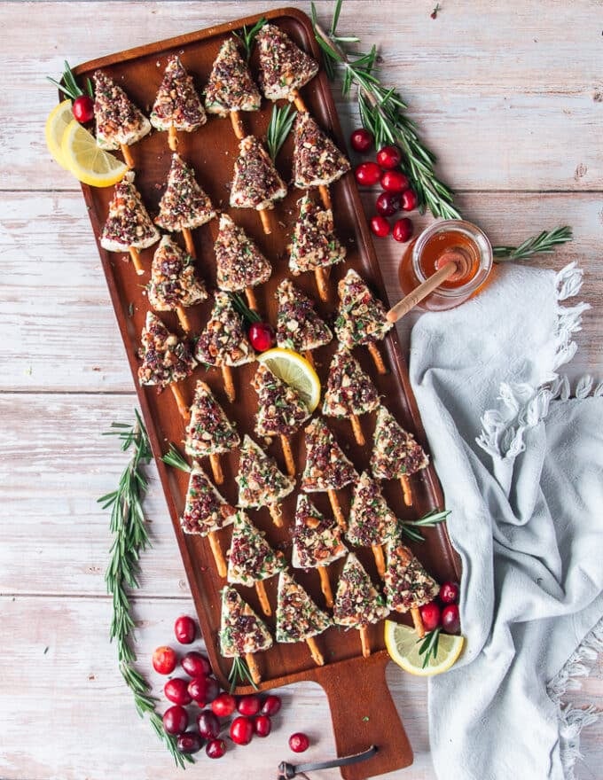 A board with small cheese Christmas trees coated in pecan, herb cranberry and lemon zest and each has a pretzel stick as a base. The board is surrounded by fresh cranberries, rosemary, lemon slices and a tea towel and looks very festive