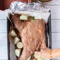 Onions and whole garlic head added around the lamb along with the whole spices in the roasting pan with the marinated lamb shoulder in the middle