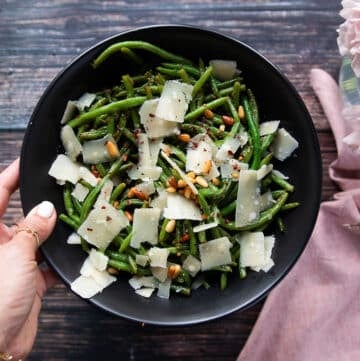 A hand holding a plate of perfect sautéed green beans. Showing the crisp tender green beans on a plate with shaved parmesan cheese, some nuts, bits of garlic and lemon zest