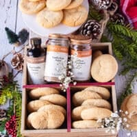 A large cookie box filled with sugar cookies and some jars of vanilla extract and vanilla bean sugar surrounded by pine cones, some flowers and a plate of more sugar cookies