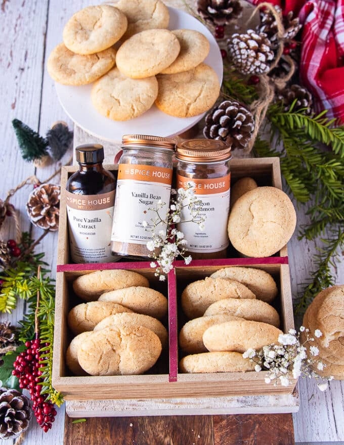 A large cookie box filled with sugar cookies and some jars of vanilla extract and vanilla bean sugar surrounded by pine cones, some flowers and a plate of more sugar cookies 