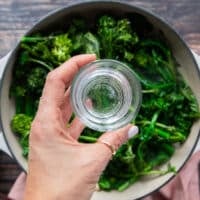 A hand holding a small glass of water over the skillet pouring it in to cook the rapini