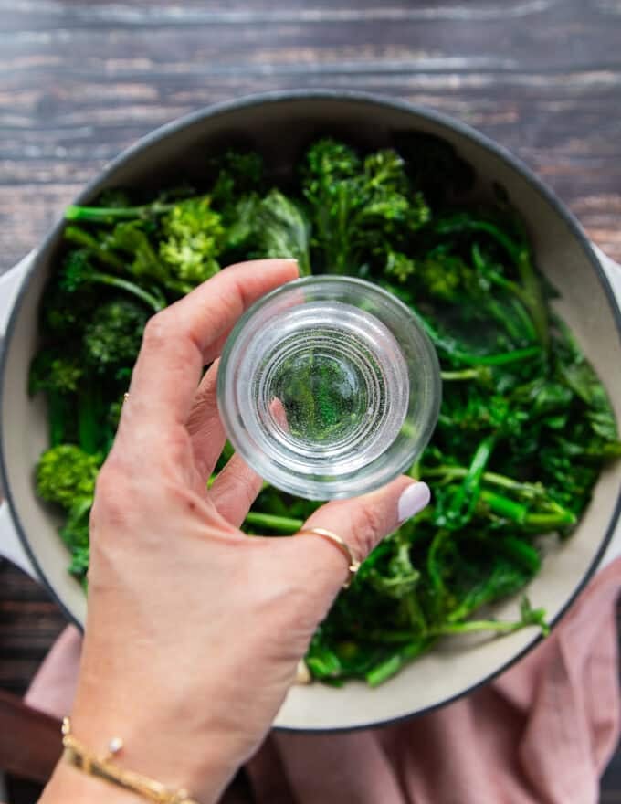 A hand holding a small glass of water over the skillet pouring it in to cook the rapini