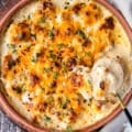 A spoon showing off the creamy cauliflower casserole showing the florets, and the creamy cheese sauce around it in a casserole dish