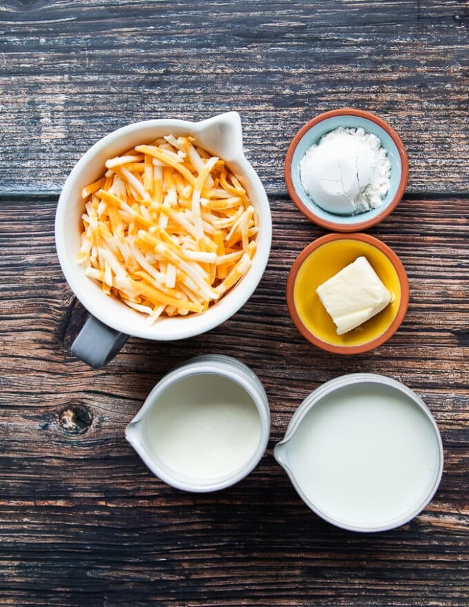 bechamel sauce ingredients on a wooden board including cups of milk, a cup of cream, a bowl of cheese, a small bowl of butter and small bowl of flour 