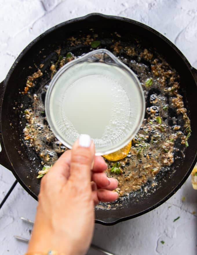 A hand holding a cup of pasta water and pouring it into the skillet to make the carb pasta sauce