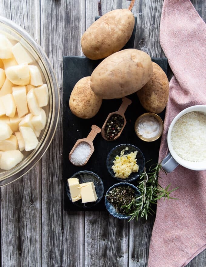 crispy potatoes ingredients on a marble including idaho potatoes, some baking soda, some water, some fresh garlic minced in a bowl, some rosemary sprigs, some grated parmesan in a bowl and a bottle of avocado oil 