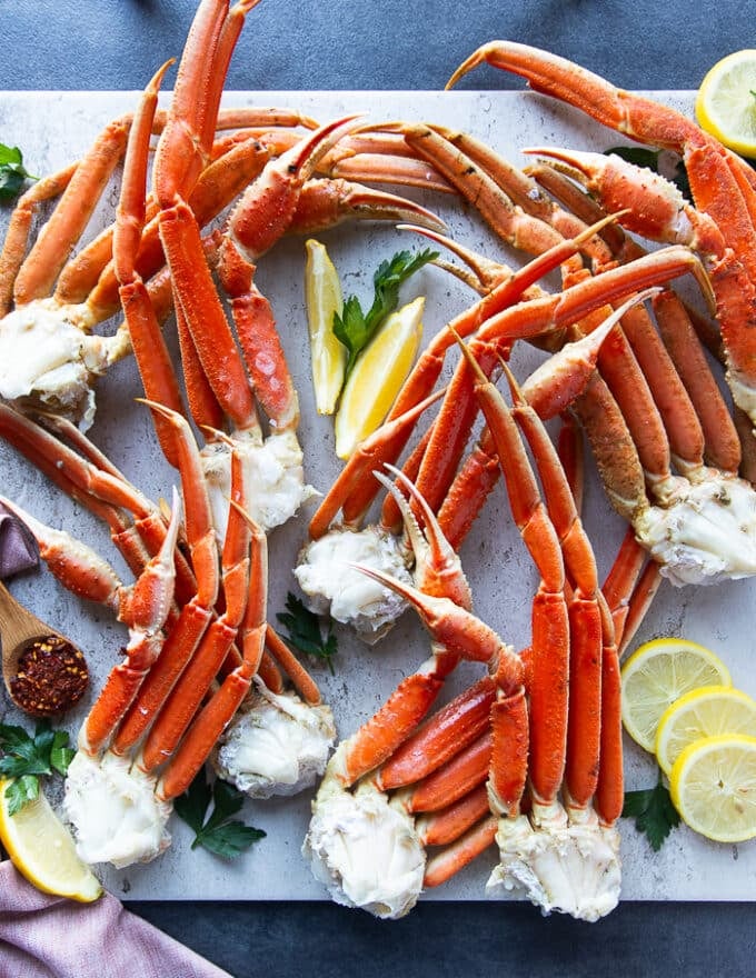 A variety of crab legs on a white marble surrounded by a bowl od old bay seasoning, some lemon slices and fresh parsley