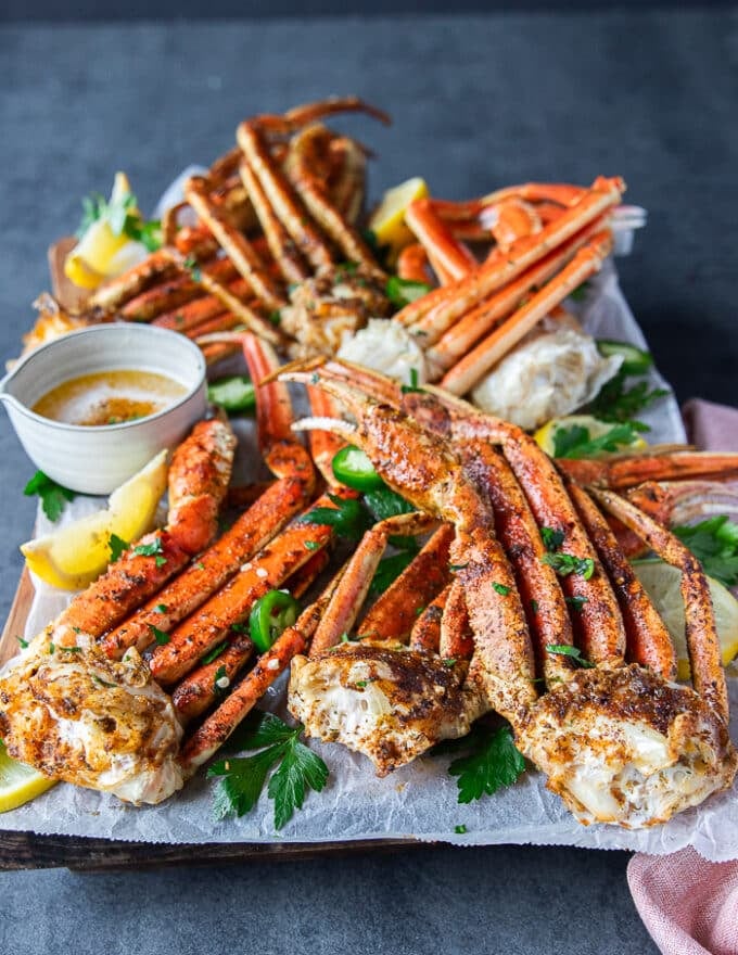 side view of a plate of cooked crab legs 4 different ways on a board surrounde dby melted butter in a bowl and lemon wedges