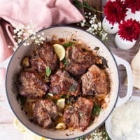 A skillet with perfectly cooked lamb chops with fresh herbs, whole garlic cloves and lemon slices