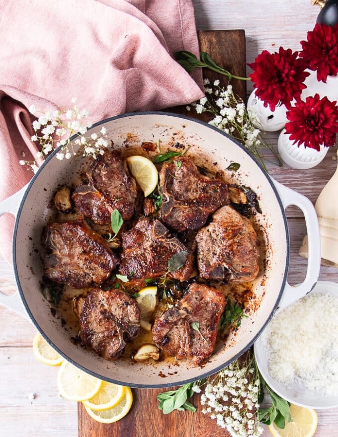 A skillet with perfectly cooked lamb chops with fresh herbs, whole garlic cloves and lemon slices 