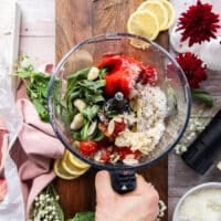 A food processor with all the romesco sauce ingredients added in one go including roasted red peppers, cherry tomatoes, almonds, lemon juice, fresh basil, olive oil, garlic, parmesan cheese and lemon juice